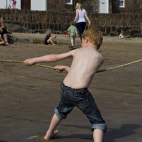 A young boy, barefoot and shirtless in denim shorts, is playing tug of war on a sandy beach in Scotland, showcasing a moment of effort and playfulness under the sun with people in the