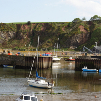 Tranquil harbor with boats at low tide, nestled against a striking cliffside under a bright blue sky in Scotland.