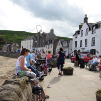 Locals and visitors enjoy a sunny day by the harbor, relaxing and socializing on the stone wall, while traditional buildings line the street behind them in a quaint Scottish coastal village, exemplifying Scotland tourism