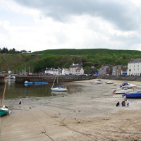 A peaceful coastal village with moored boats and visitors enjoying the tranquil beach during low tide, set against a backdrop of green hills and overcast skies. This sight embodies the essence of Scottish culture.