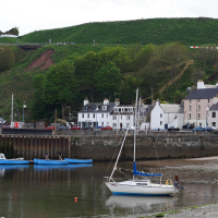 A serene harbor scene in the Scottish Highlands with moored boats, a low tide exposing muddy shores, quaint buildings by the waterfront, and a lush green hill with a path in the background under a cloudy