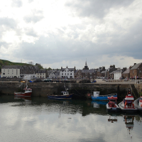 A tranquil harbor in Scotland, with moored boats reflecting in the calm water, under an overcast sky, with a picturesque row of buildings lining the waterfront.