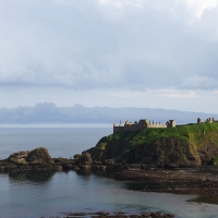 Ruined fortress perched on a green cliff in Scotland, overlooking a calm sea against a backdrop of moody skies, with scattered sunlight illuminating the historic structure and serene waters.