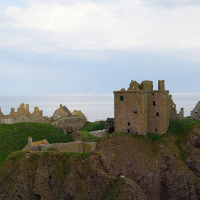 Ancient ruins of a seaside castle in Scotland, perched atop rugged cliffs, with partially intact towers and crumbling walls, under a soft blue sky scattered with birds in flight.