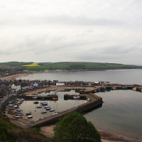 A tranquil harbor in Scotland on a cloudy day, with small boats moored in calm water, flanked by a quaint coastal village and rolling hills in the background, presenting a serene marine landscape.