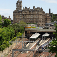 A multi-level train station nestled between lush greenery in Scotland, with rail tracks in the foreground, under a clear blue sky, with a historic baroque-style building dominating the background.