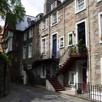 Charming cobbled lane in Edinburgh's historic district, a prime destination for tourism, featuring stone townhouses with colorful doors, detailed ironwork, and lush window boxes under a canopy of greenery.