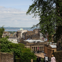 A scenic view from an elevated street in Edinburgh, Scotland, capturing pedestrians enjoying a sunny day, with the city's historic buildings in the background and the Firth of Forth visible in the distance.