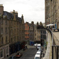 Historic multistory buildings line an old cobblestone street bustling with activity, showcasing urban life in a traditional Scottish city.