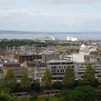 A panoramic view of Edinburgh's varied architecture, showcasing Scotland's historic buildings in the foreground, modern developments amidst green spaces, and the Firth of Forth visible in the distance under a partly cloudy sky