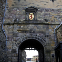 A historical stone gateway featuring a portcullis, with the crest of arms above, leading to an old fortress, contrasting modern visitors with its ancient structure, epitomizes Scottish tourism.