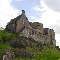 A historic stone building perched on a rocky hillside in the Scottish Highlands under a cloudy sky, with a cobblestone path leading up to it, a traditional red post box standing prominently in the