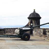 An antique cannon perched atop an old fortress wall in Scotland, with a panoramic view stretching to the hazy horizon under a partly cloudy sky.