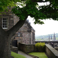 An old stone building with a leaded window peeks out from behind lush green tree branches, overlooking a historical courtyard in Scotland, with a distant view of city spires under a cloudy sky.