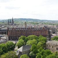 Panoramic view of a historic cityscape in Scotland featuring classic architecture, with church spires and heritage buildings nestled among verdant trees under a cloudy sky.