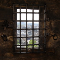 A heavy, black, barred window set in a thick stone wall overlooks a cityscape in Scotland, juxtaposing the confinement of the historic structure with the expansive view of the urban horizon beyond.