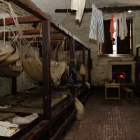 A historic marine barracks room in the Highlands with hammocks for sleeping, a stone fireplace lit for warmth, personal items scattered, and laundry hanging overhead, reflecting the austere living conditions of sailors in the