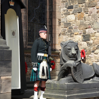 A ceremonial guard in traditional Scottish attire, including a kilt, stands at attention beside a stone lion sculpture, against the backdrop of a historic stone building in Scotland.