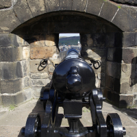 A historical cannon on display, poised at an opening in a Scottish stone fortification, with a cityscape visible in the background beneath a clear blue sky.