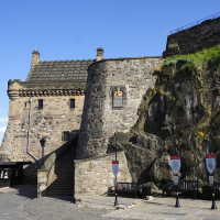 A stone building with a slate roof at the base of a rocky cliff in Scotland, adorned with heraldic shields, under a clear blue sky, likely a historic fortress or castle gatehouse.