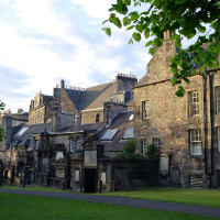 Historic stone buildings with slate roofs stand majestically under a clear blue sky, flanked by lush green trees in a serene park-like setting, showcasing classic Scottish architecture.