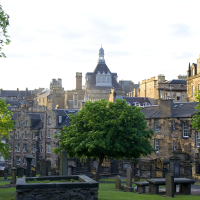 A serene graveyard with historical tombstones amid lush greenery, framed by classic stone buildings under a clear blue sky, capturing the tranquil beauty and architectural heritage of Scotland.