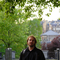 A contemplative man stands in a serene cemetery in Scotland, with lush greenery and historic buildings softly blurred in the background, evoking a sense of tranquility amidst the vestiges of the past.