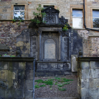 An aged, ornate stone monument with inscriptions, set against a weathered brick building in Scotland, under the tranquil shade of a tree, with greenery sprouting from its base.