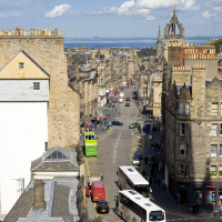 A bustling street in a historic city in Scotland, lined with old stone buildings under a clear blue sky, as buses and cars navigate the thoroughfare.