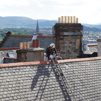 A worker in safety gear performs maintenance on a steep slate roof in Scotland, with a backdrop of rolling hills and city buildings under a partly cloudy sky.