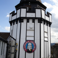 A unique, octagonal, black-and-white camera obscura tower in Scotland, with a sign featuring its name, juxtaposed against a partly cloudy sky.