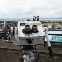 A coin-operated binocular viewer overlooking a panoramic cityscape under partly cloudy skies, offering tourists a closer look at the urban horizon in Scotland.
