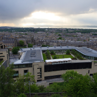 An aerial view of a modern urban landscape in Scotland at dusk, showcasing a mix of contemporary architecture and traditional buildings against a backdrop of a dramatic sky pierced by rays of sunlight.