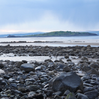 A tranquil rocky beach in Scotland at low tide with scattered boulders and puddles reflecting the sky, overlooked by distant hills beneath a sky where rain is visibly falling in the background.