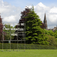 An art installation displaying the phrase "there will be no miracles here" stands in a lush, green park in Scotland, with towering spires visible in the distance under a partly cloudy sky, highlighting a