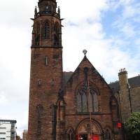 An imposing gothic revival church with a tall spire stands under a partly cloudy sky, featuring arched windows and a rich red-brown facade, embodying Scottish culture, while pedestrians pass by on