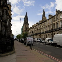 A picturesque street in a Scottish city, showcasing classical architecture with spire-topped towers in the distance, a clear blue sky overhead, and vehicles parked along the sidewalk.