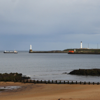 A serene coastal scene in Scotland featuring a beaconing lighthouse accompanied by a smaller structure, under a cloudy sky, flanked by a cargo ship and rocky shore, viewed across tranquil waters from a sandy