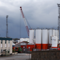 A gloomy sky looms over a Scottish industrial harbor scene, featuring towering white storage tanks, a large red and white crane, and modest buildings, with a hint of maritime activity in the background.