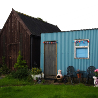 A quaint blue corrugated metal shack adorned with colorful curtains and flower decorations stands next to a larger wooden barn, surrounded by a charming garden with potted plants, outdoor chairs, and an overcast