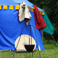 A blue tent with yellow trim in a grassy area has blankets and garments hanging on the side. A round wicker mat lies on the ground, and a black pot hangs from a tripod stand in the foreground. Trees and greenery are visible in the background, evoking memories of Semigallian Days Festival 2009.