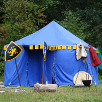 A blue medieval-style tent with yellow and black trim stands in a grassy area with a forest in the background. In front of the tent are two shields, wooden logs, and a black cauldron on a tripod. Garments and items like a shield and furs are draped over the tent's entrance, reminiscent of Semigallian days.