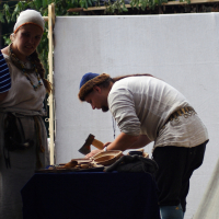 Two individuals in medieval attire are engaging in traditional crafts during the Semigallian Days of 2009. A woman, wearing a white headscarf and adorned with necklaces, stands on the left. A man in a blue cap works with a small axe on a wooden block to the right. A table with wooden bowls and tools sits in the foreground.