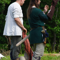 Two men in medieval attire stand outdoors at Semigallian Days '09. The man on the left has a shaved head and wears a white tunic with brown pants, holding part of a dragon costume. The man on the right sports a fur hat, green tunic, and light pants, adjusting a sleeve of the dragon costume amidst lush greenery.