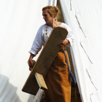 A woman dressed in historical clothing, consisting of a white blouse and a long brown skirt, stands in front of a white tent at the 2009 Semigallian Days festival. She holds a large wooden object, possibly a tool or part of a structure. Her hair is tied back, and she appears to be engaged in an outdoor activity or event.