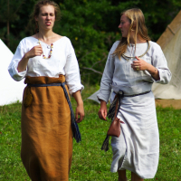 During the Semigallian Days in 2009, two women walk barefoot on grass, dressed in traditional medieval attire. One wears a white blouse and brown skirt with a tied blue belt, while the other dons a white dress with a blue sash and a small pouch. Both have long hair, and tents can be seen in the background amidst greenery.