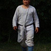 A person, dressed in medieval-style clothing, is walking outdoors during the 2009 Semigallian Days. They wear a loose, long-sleeved white tunic with a belt, white breeches, and blue leg wraps. The background is dark green foliage. The lighting suggests a natural setting with a mix of sunlight and shadow.