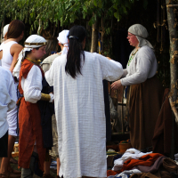 A group of people, some in historical or medieval attire, are at an outdoor event under a leafy canopy. One person in a long white robe and bare feet stands with their back to the camera. Others wear various period costumes, including head coverings and tunics, reminiscent of the 2009 Semigallian festival.