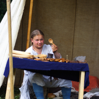 At a Semigallian festival, a person with long hair, dressed in historical or traditional clothing, sits under a beige canvas tent. They are at a wooden table covered with a blue cloth, displaying wooden utensils. The person holds up a wooden spoon, examining it closely. The grassy ground and other items are visible around them.