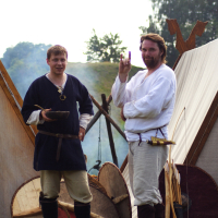 Two men dressed in medieval-style clothing stand in front of canvas tents at the Semigallian Days cultural event. The man on the left, in a navy tunic and beige trousers, holds a wooden bowl. The man on the right, in a white tunic and trousers, holds up a small purple object. Shields rest against the tents behind them.